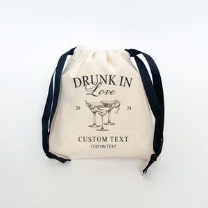 May include: A white drawstring bag with black drawstrings. The bag has a black print that says "Drunk in Love" with a drawing of three martini glasses. The text "Custom Text" is printed below the drawing.