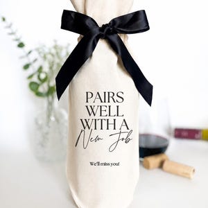 May include: A cream-colored fabric wine bottle gift bag featuring a black satin ribbon tied at the top. The bag is printed with the black text "PAIRS WELL WITH A New Job" and "We'll miss you!" in a combination of serif and script fonts.