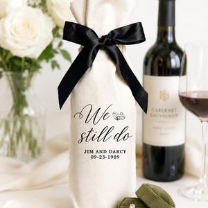 Pode incluir: Um saco de presente de tecido cor de creme para garrafa de vinho com um laço de fita de cetim preta no topo. O saco está impresso com o texto "We still do" acima dos nomes "JIM AND DARCY" e a data "09-23-1989" em fonte preta.