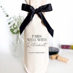 Op de afbeelding: Een crèmekleurige stoffen geschenkzak voor wijnflessen met een zwarte satijnen strik aan de bovenkant. De zak is bedrukt met de zwarte tekst PAIRS WELL WITH Retirement en de kleinere tekst No more Sunday scaries in het midden aan de voorzijde.