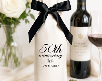 May include: A beige fabric wine bottle gift bag tied with a black satin ribbon. The bag features black printed text that reads 50th anniversary, a graphic of two interlocking rings, and the names TOM & KAREN. An olive green velvet ring box is nearby.