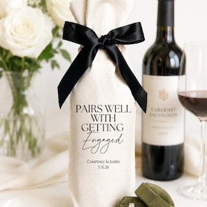 Puede incluir: Una bolsa de regalo de tela color crema para botella de vino, atada con una cinta de satén negra. La bolsa tiene el texto impreso en negro: PAIRS WELL WITH GETTING Engaged, Courtney & Justin, 5.31.26. Un joyero de terciopelo verde oliva está delante.
