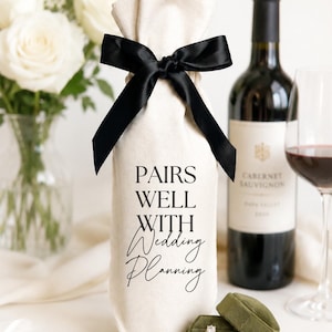May include: A cream-colored fabric wine bottle gift bag featuring a black satin ribbon tie at the top and the black printed text PAIRS WELL WITH Wedding Planning. An olive green velvet ring box with a gold ring is placed in front.