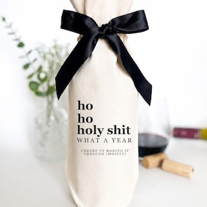 Könnte beinhalten: Ein cremefarbener Stoffbeutel für Weinflaschen, der mit einer schwarzen Satinschleife gebunden ist. Der Beutel ist mit schwarzem Text bedruckt: ho ho holy shit WHAT A YEAR CHEERS TO MAKING IT THROUGH (MOSTLY). Der Beutel ist zylindrisch geformt.