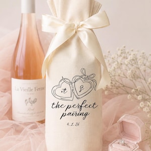 May include: A cream-colored fabric wine bottle gift bag tied with a matching satin ribbon. The bag features a black line-art graphic of two connected hearts with the letters A and S, the text "the perfect pairing," and the date 4.1.26.