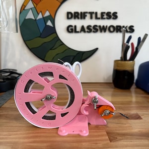 May include: A pink 3D printed tape dispenser with a white tape roll and a black label that reads "Driftless Glassworks". The dispenser has a small orange wheel and a metal blade.