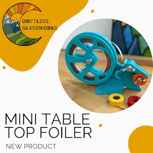 May include: A blue and brown tabletop foiling machine with a white wheel and a yellow, orange, and red spool. The machine is on a wooden surface. The text "Driftless Glassworks" is visible in the image.  "Mini Table Top Foiler" and "New Product" are also visible in the image.
