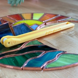 May include: A yellow plastic tool with the text "Driftless Treasures" and "1/4" printed on it. The tool is used for stained glass projects and is sitting on top of a stained glass piece that depicts a sunset with a mountain range and a wavy ocean.