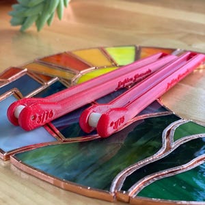 May include: Two bright red 3D-printed plastic tools with white rollers, each marked with the text 3/16, resting on a stained glass piece featuring green, yellow, and orange segments joined by copper foil lines.