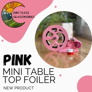 May include: Pink mini table top foiler, a new product from Driftless Glassworks. The foiler is a small, pink, hand-cranked device with a spool of foil.