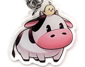 May include: A white and black cow keychain with a small yellow chick on its head. The keychain is made of clear acrylic and has a silver metal ring.