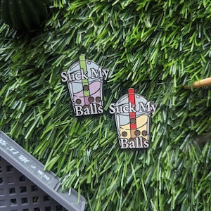 K&ouml;nnte beinhalten: Zwei Emaille-Pins in Form von Bubble-Tea-Bechern mit dem Text "Suck My Balls" auf jedem. Die Bubble-Tea-Becher sind mit bunten Boba-Perlen gef&uuml;llt.