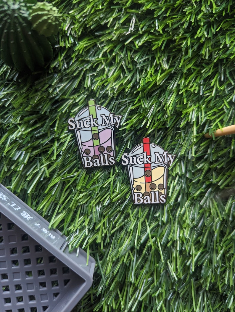 K&ouml;nnte beinhalten: Zwei Emaille-Pins in Form von Bubble-Tea-Bechern mit dem Text "Suck My Balls" auf jedem. Die Bubble-Tea-Becher sind mit bunten Boba-Perlen gef&uuml;llt.