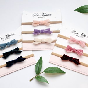 May include: Three sets of velvet hair bows on nude elastic headbands. Each set includes three bows in different colours: pink, purple, and cream; blue, teal, and navy; and burgundy, pink, and cream. The bows are arranged on white cards with the text "Mon Chaton Boutique".