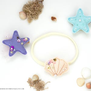 May include: A set of three felt hair clips featuring a purple starfish with pink and purple sequins, a light blue starfish with white sequins, and a pink seashell with white sequins. The clips are displayed on a white background with seashells and a headband.