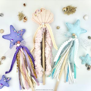 May include: Three handmade wands with a whimsical sea theme. Each wand features a different topper: a purple starfish, a blue starfish, and a pink seashell. The wands are decorated with colourful ribbons, tulle, and pom-poms.