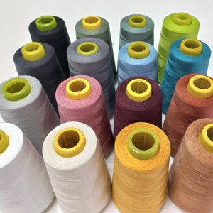 Könnte beinhalten: Sortiment von Garnrollen in verschiedenen Farben, darunter Weiß, Grau, Braun, Rot, Blau, Grün und Gelb. Die Rollen sind in einem Kreis angeordnet.