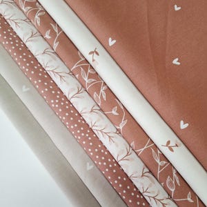 Könnte beinhalten: Eine Sammlung von Stoffmustern in verschiedenen Mustern und Farben. Die Stoffe umfassen unifarbene Beige-, Punkt-, Blumen- und Herzmuster in Beige-, Weiß- und Terrakotta-Tönen. Die Muster sind diagonal angeordnet und zeigen die verschiedenen Texturen und Muster.
