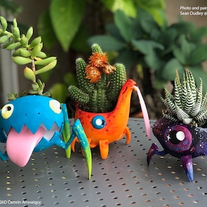 May include: Three 3D printed planters shaped like cartoon monsters, each with a different colour scheme and design. The planters are holding succulents, and the text "MonsterPots 5IX26D Camrin Arreseigor" is visible at the bottom of the image.