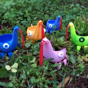 May include: Five colorful monster-shaped planters with long tongues, each holding a small plant. The planters are made of 3D printed plastic and have a whimsical design. The text "Monster Pots" is visible at the bottom of the image.