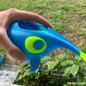 May include: A blue 3D printed toy shaped like a bird with a yellow eye and a long beak. The toy is being held in a person's hand.