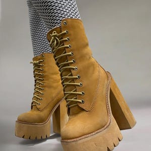 May include: Tan platform boots with thick, lug soles and high block heels. The boots have a lace-up design with tan laces and a padded collar. The boots are made of a suede-like material and have a rugged, outdoorsy look.