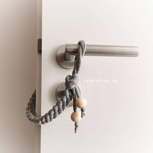 Könnte beinhalten: Grauer geflochtener Türstopper aus Stoff mit zwei Holzperlen und einem Knoten am Ende. Der Türstopper ist an einem Türgriff befestigt.