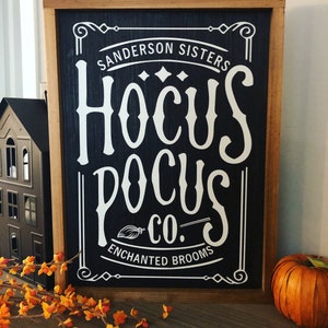 May include: A black and white sign with a wooden frame. The sign reads "Sanderson Sisters Hocus Pocus Co. Enchanted Brooms".