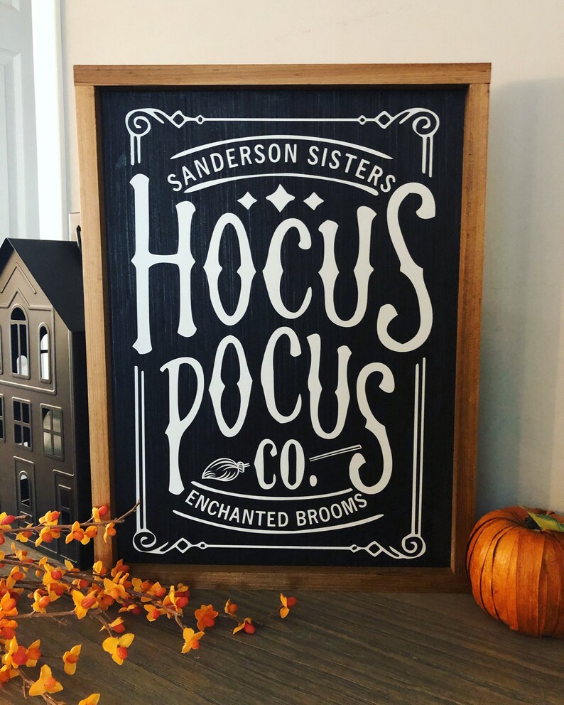 May include: A black and white sign with a wooden frame. The sign reads "Sanderson Sisters Hocus Pocus Co. Enchanted Brooms".