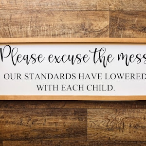 May include: A white wooden sign with a brown frame reads "Please excuse the mess our standards have lowered with each child."