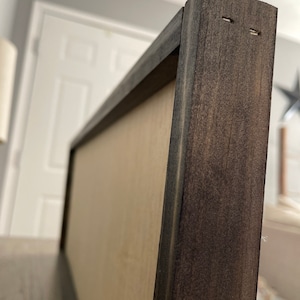 May include: Close-up of a dark brown wooden frame with a light brown backing. The frame has two small holes in the top corner.