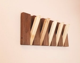 Può includere: Un appendiabiti da parete in legno con sei ganci. I ganci sono in legno chiaro e sono inclinati verso l'alto. L'appendiabiti è in legno scuro e ha una finitura in legno naturale.