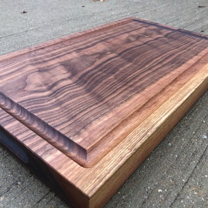 May include: A large, rectangular walnut cutting board with a raised edge and a small handle. The board has a dark brown color and a natural wood grain.