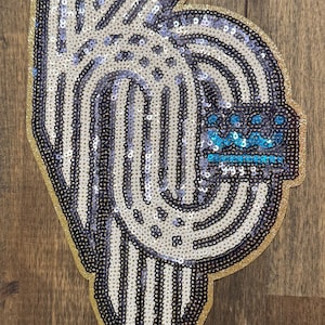 May include: A sequined patch in the shape of the letters "KC" with a gold glitter border. The design features alternating vertical stripes of white and dark blue sequins. A small section of blue sequins is at the top.