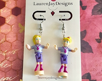 May include: A pair of earrings featuring miniature plastic figures of girls wearing purple dresses and pink shoes. The earrings are displayed on a white card with the text "LaurenJayDesigns" and "laurenjaydesigns.com".