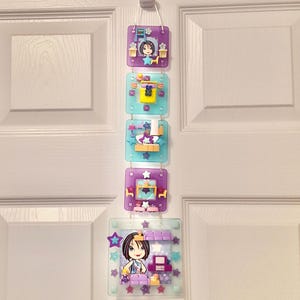 May include: A door hanger with five clear, square panels, each displaying a colourful anime-style illustration. The panels are linked and suspended from a white door. Each panel features a different scene with stars and a character.