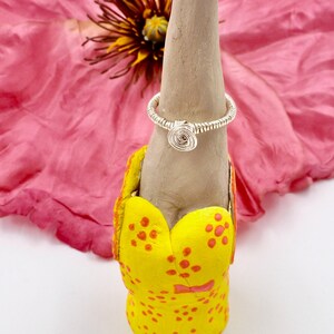 May include: A silver wire ring with a spiral design is displayed on a grey ring holder. The ring holder is set on a yellow base with red polka dots. A large pink flower is in the background.