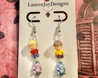 May include: A pair of dangle earrings featuring a cartoon boy and a teddy bear charm. The boy is wearing a red shirt and blue pants. The teddy bear is purple and blue. The earrings are on a white card with the text "LaurenJayDesigns" and "laurenjaydesigns.com".
