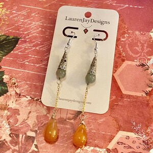 May include: A pair of dangle earrings with a green stone and a gold chain. The earrings are on a white card with the text "LaurenJayDesigns" and "laurenjaydesigns.com".
