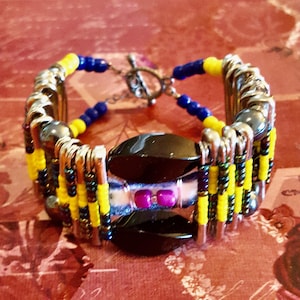 Op de afbeelding: Een kleurrijke armband gemaakt met veiligheidsspelden, kralen en een zwarte steen. De armband is geel, blauw en zwart met een paarse accent.