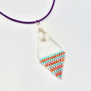 Op de afbeelding: Een zilveren draad driehoekige hanger ketting met een turquoise en oranje kralen ontwerp. De ketting is geregen op een paars koord.