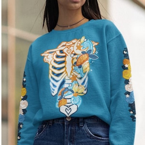 May include: Two women wearing blue and orange sweatshirts with a skeleton and floral design. The design features a ribcage with flowers, seashells, and a heart.