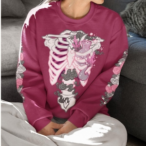 Könnte beinhalten: Ein weinrotes Sweatshirt mit einem weißen Brustkorb-Design und rosa und grauen floralen Akzenten. Die Ärmel haben ein Blumenmuster. Das Sweatshirt wird von einer Person getragen, die auf einem Sofa sitzt.