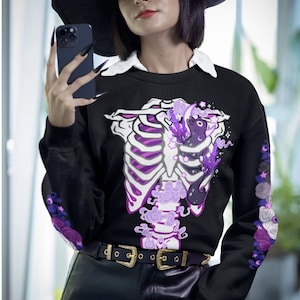 May include: Two purple sweatshirts with a skeleton and floral design. One sweatshirt is black and the other is purple. Both sweatshirts have the text "Skeleton Floral" on them.