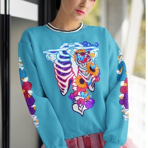 May include: Two women wearing blue and pink sweatshirts with a colorful graphic of a tiger skeleton with flowers and butterflies. The sweatshirts have long sleeves and a crew neck.