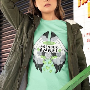 May include: A light green t-shirt with the words "AGENDER ANGEL" and a graphic of angel wings, flowers, and leaves. The person is wearing a green jacket and blue jeans.
