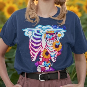 May include: A navy blue t-shirt with a graphic of a tiger inside a ribcage with sunflowers and butterflies. The tiger has blue eyes and a pink nose.