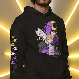 Könnte beinhalten: Ein schwarzer Hoodie mit einem Aufdruck einer Gruppe von Katzen, die als Geister und Hexen verkleidet sind. Der Aufdruck enthält einen Grabstein mit der Aufschrift "RIP". Der Hoodie hat lila und gelbe Pfotenabdrücke auf den Ärmeln.