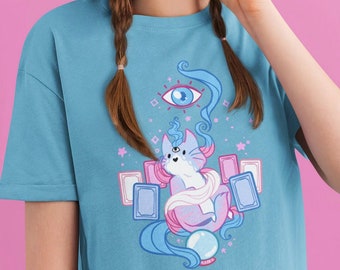 May include: A light blue t-shirt with a graphic of a cartoon cat with a third eye, surrounded by tarot cards and a crystal ball. The cat is wearing a white scarf and has blue hair. The background is a light blue with stars and other celestial symbols.