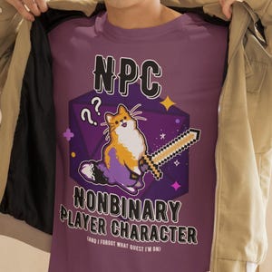May include: A purple t-shirt with the text "NPC NONBINARY PLAYER CHARACTER" and a cartoon cat holding a pixelated sword. The shirt also has the text "(AND I FORGOT WHAT QUEST I'M ON)".